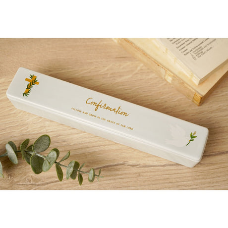 White ceramic rectangular Confirmation certificate keepsake box with gold cross and dove motifs, displayed on a wooden surface beside eucalyptus leaves and a book.