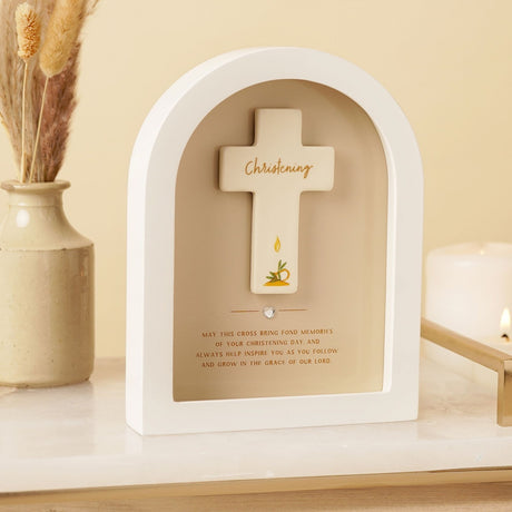 White arched wooden Christening plaque with cream cross, gold Christening script, and candle motif, displayed on a shelf beside a dried flower vase and lit candle.