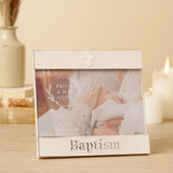White enamel metal photo frame with silver trim, raised "Baptism" lettering and a small cross, shown on a table with candles in a lifestyle setting.