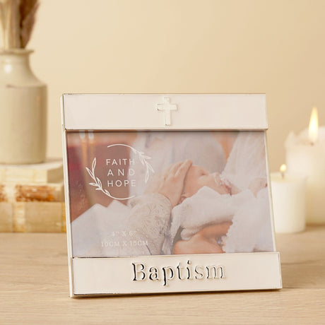 White enamel metal photo frame with silver trim, raised "Baptism" lettering and a small cross, shown on a table with candles in a lifestyle setting.