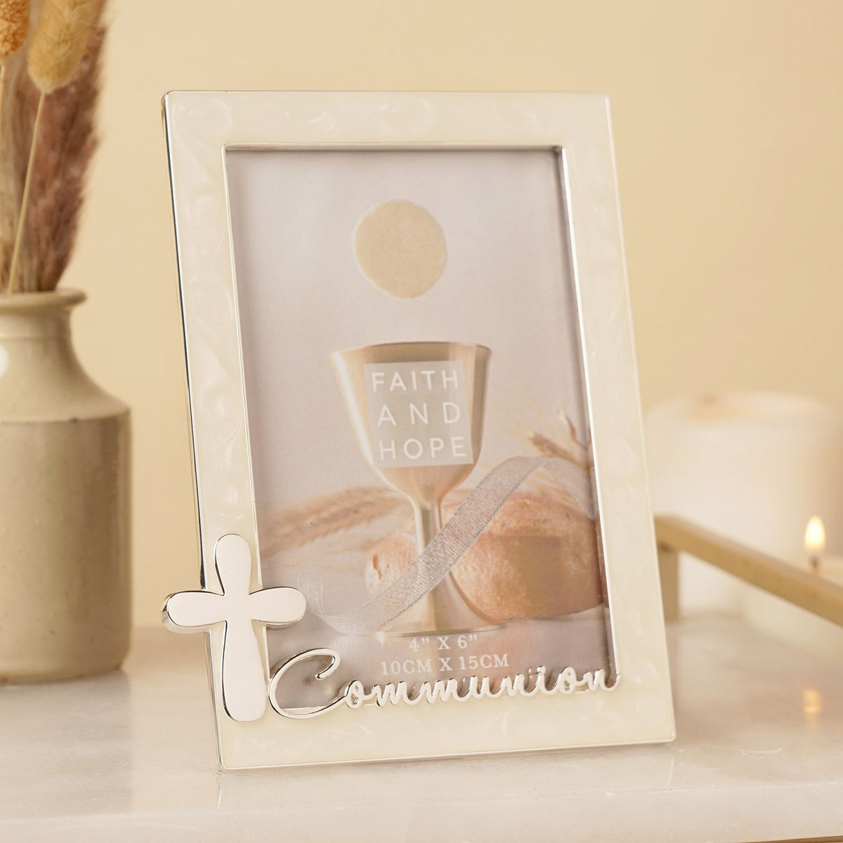 Cream enamel metal photo frame with silver trim, raised silver cross, and "Communion" script, holding a Faith and Hope sample print, displayed on a shelf beside a vase and candle.