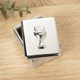 Rectangular silver metal keepsake box with fitted lift-off lid, featuring a raised chalice and IHS host design, shown on a wooden surface with a leaf sprig nearby.