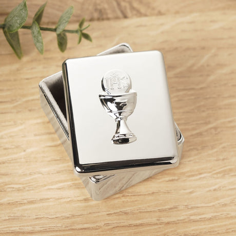 Rectangular silver metal keepsake box with fitted lift-off lid, featuring a raised chalice and IHS host design, shown on a wooden surface with a leaf sprig nearby.
