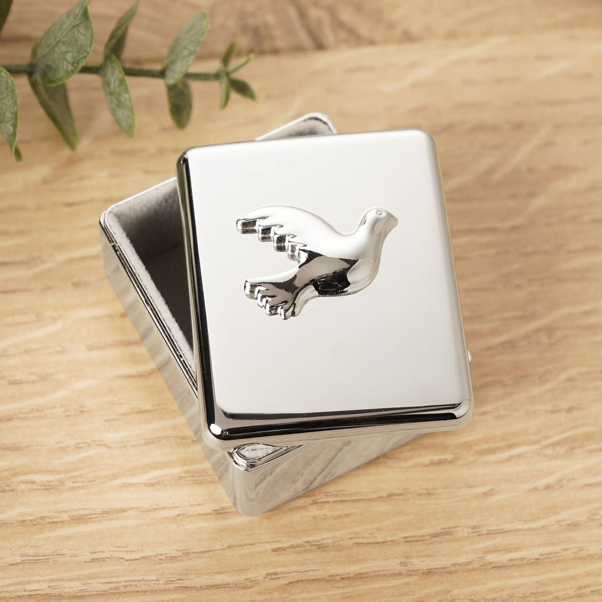 Rectangular Dove Confirmation metal keepsake box in polished silver, lid featuring a raised dove in flight, shown on a wooden surface with greenery nearby.