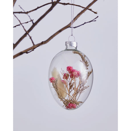 Fintry Decorative Egg, a clear glass hanging ornament with silver cap and loop, filled with pink flower heads, brown sprigs and a cream grass plume, shown against a bare branch on a light background.