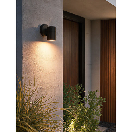 Firvette Outdoor Wall Light in black, aluminium fluted cylindrical head on a round wall plate, mounted beside a wooden front door and illuminating the render wall below.