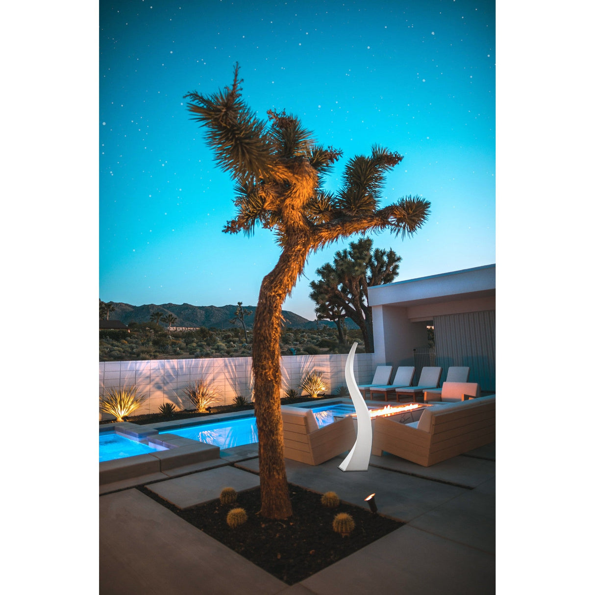 At dusk, a modern patio is lit by the Flame 5 Light Outdoor Floor Lamp in Opal White, illuminating a Joshua tree beside a fire pit with white lounge chairs, near a pool and mountains under a star-filled sky.