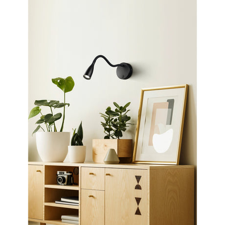 Flarmora LED Wall Light in black, with a round steel backplate and flexible gooseneck arm, mounted above a wooden sideboard with plants and framed art in a living room.