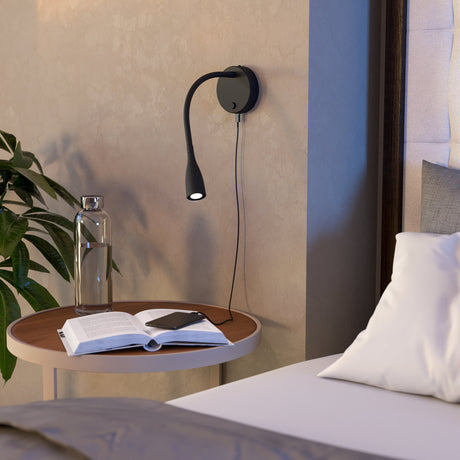 Flarmora LED wall light in black with round steel plate, flexible gooseneck arm and tapered head, shown mounted above a bedside table with a USB cable visible.