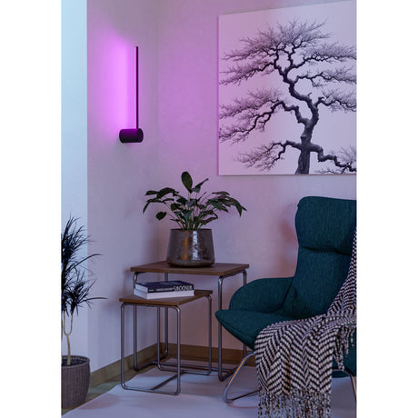 Flatron Smart Colour Changing LED Wall Light in matt black, mounted vertically glowing pink, shown beside artwork in a living room with armchair and side tables.