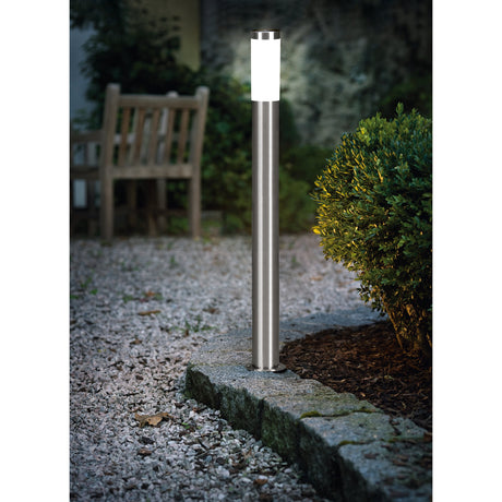 Flerbenne stainless steel bollard light illuminated on a garden gravel path, its white diffuser top glowing beside a stone border, bench and shrubs in the background.