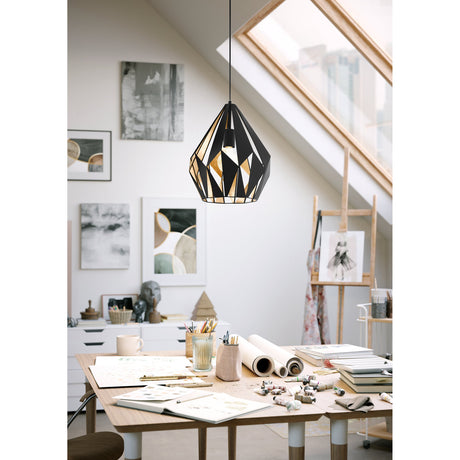 Flermanne Pendant Light in black and gold faceted steel, hung from a slim black cord above a wooden desk in a bright attic studio workspace.