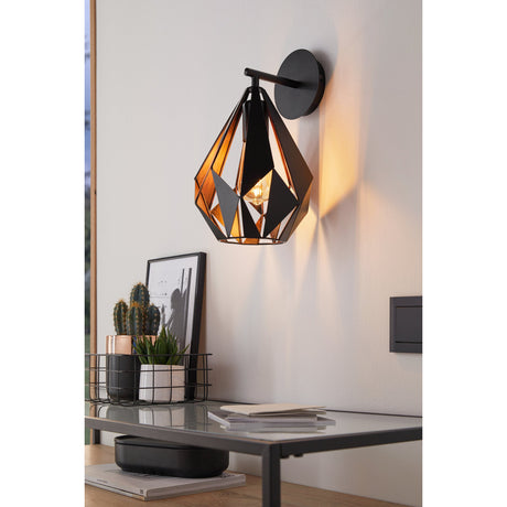 Flermanne wall light with a faceted teardrop steel shade, black outside and copper-lined inside, mounted on a round black wall plate above a living room console table.