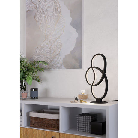 Flimarne LED Table Lamp in black, with looping aluminium and steel rings and a white diffuser, shown lit on a sideboard in a living room setting.