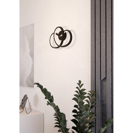 Flimarne LED Wall Light in black, with intertwining aluminium and steel loops, mounted on a light wall above a plant in a living room setting.