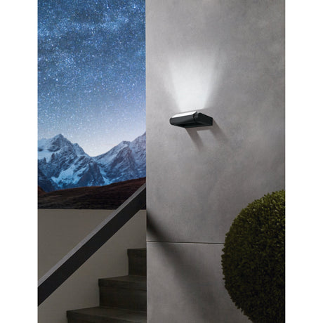 Flirmane LED Outdoor Wall Light in black aluminium, slim rectangular frame with cylindrical head, mounted on an exterior grey wall casting an upward light beam.