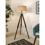 Flivelle Floor Lamp with walnut wood tripod legs, black steel joints and a taupe fabric drum shade, shown in a living room corner setting.
