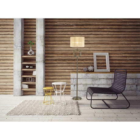 A modern living space with wooden panel walls, a Florence Round 1 Light Crystal Floor Lamp in brass with a cream shade, a black wire chair, two small round tables, a beige rug, wall shelves with decor, and empty picture frames on the floor.