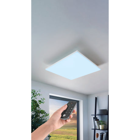 Flornorne square LED flush ceiling light in white finish mounted on a room ceiling, shown lit with a handheld remote control for colour and dimming settings.