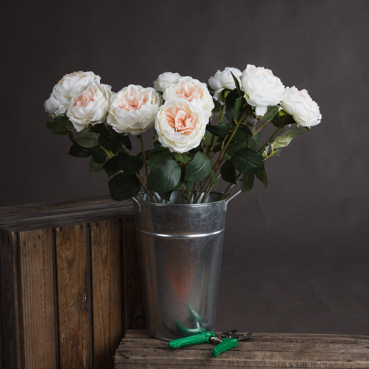 Hill Interiors Blush Garden Rose faux stems with layered blush pink and cream petals, arranged in a galvanised metal bucket beside a wooden crate.