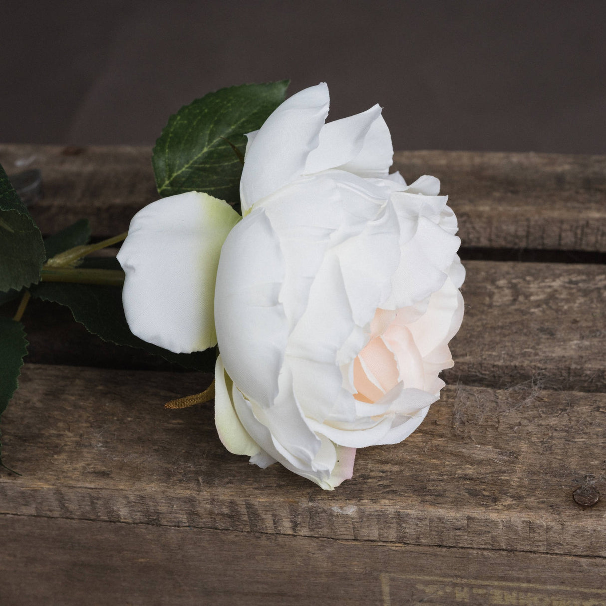 Hill Interiors Blush Garden Rose faux flower with layered white and blush pink petals on a bendable stem with green leaves, resting on rustic wood.
