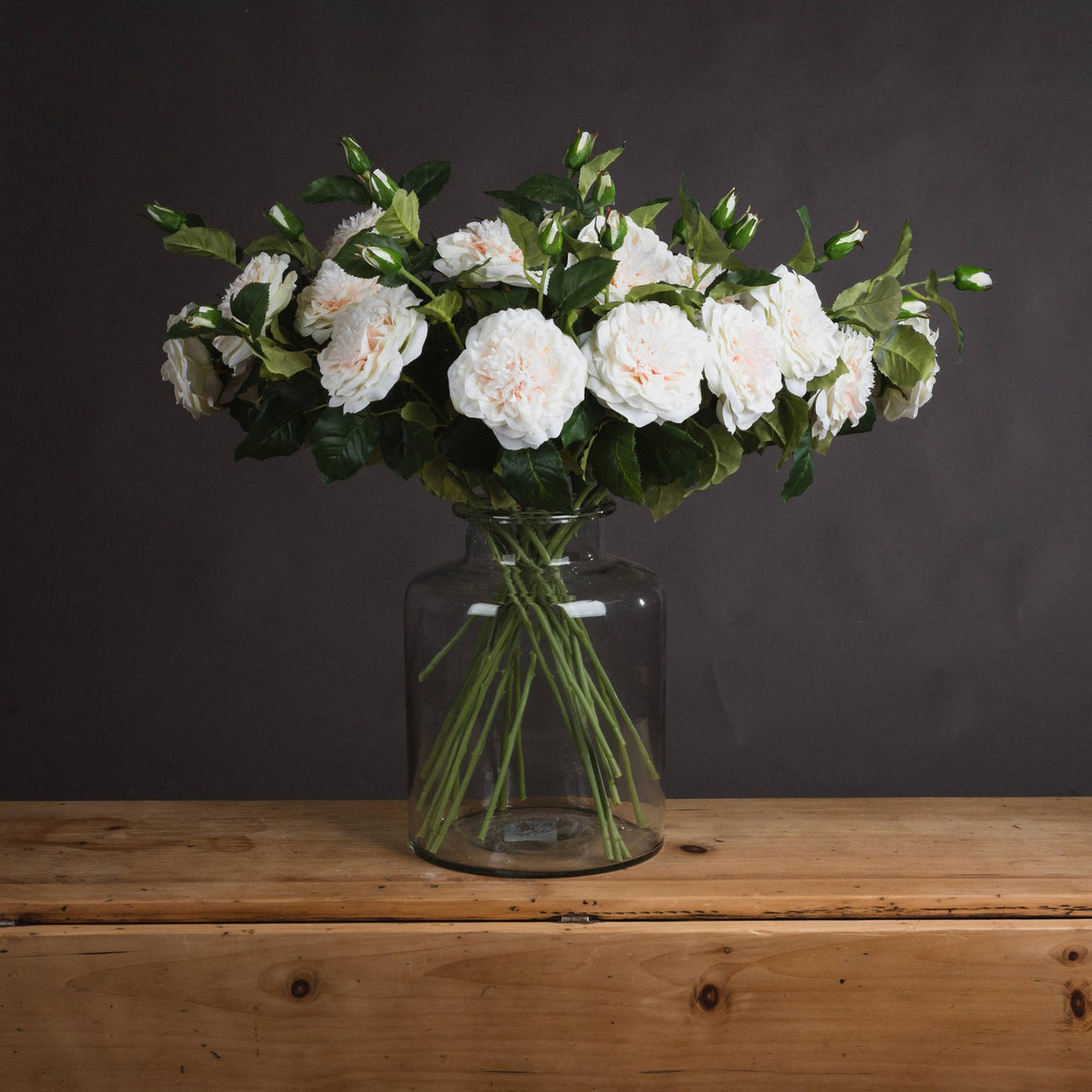 Hill Interiors White Garden Rose Spray arranged in a clear glass jar vase on a wooden surface against a dark grey background.
