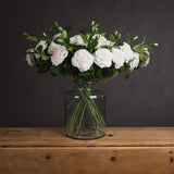 Hill Interiors White Garden Rose Spray arranged in a clear glass jar vase on a wooden surface against a dark grey background.