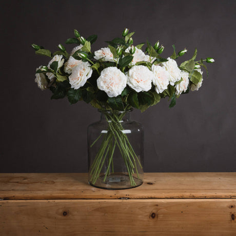 Hill Interiors White Garden Rose Spray arranged in a clear glass jar vase on a wooden surface against a dark grey background.