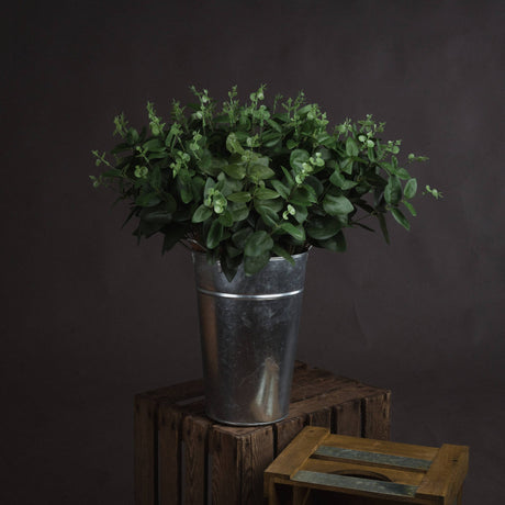 Hill Interiors Cineraria Eucalyptus Spray with muted green paired leaves arranged in a galvanised metal bucket on a wooden crate against a dark backdrop.
