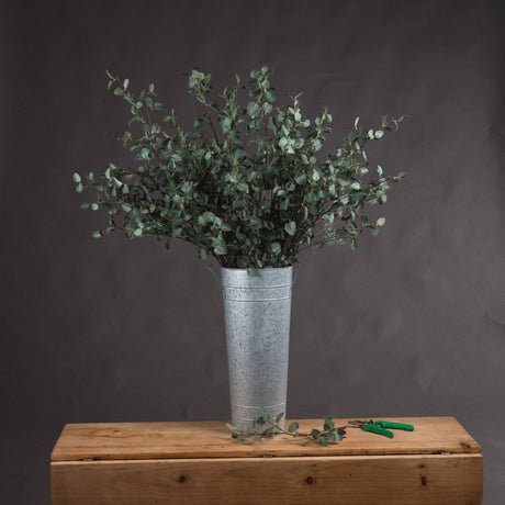 Hill Interiors Tall Eucalyptus Stems arranged in a galvanised metal florist bucket on a wooden chest, with green-handled snips beside it.