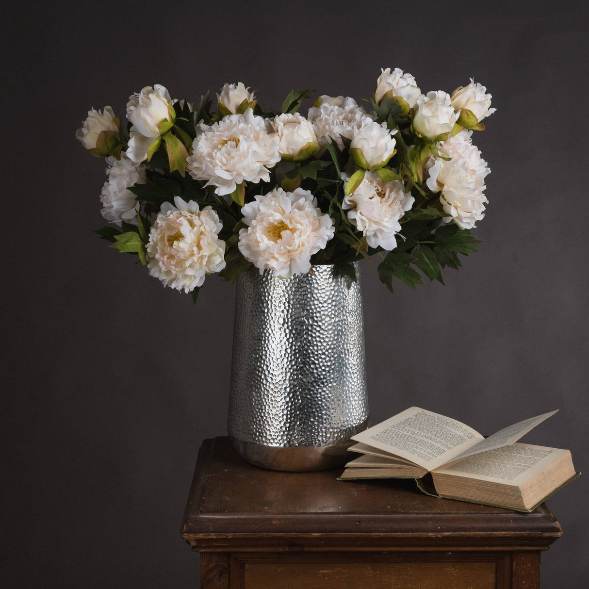 Hill Interiors White Fashion Peony faux stems arranged in a hammered silver vase on a wooden side table beside an open book against a dark grey backdrop.
