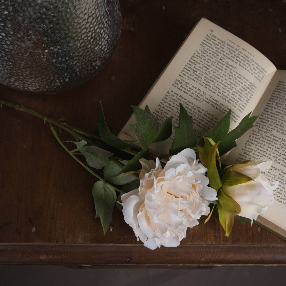 Hill Interiors White Fashion Peony faux stem with ruffled cream petals and green foliage resting on an open book beside a textured glass vase.