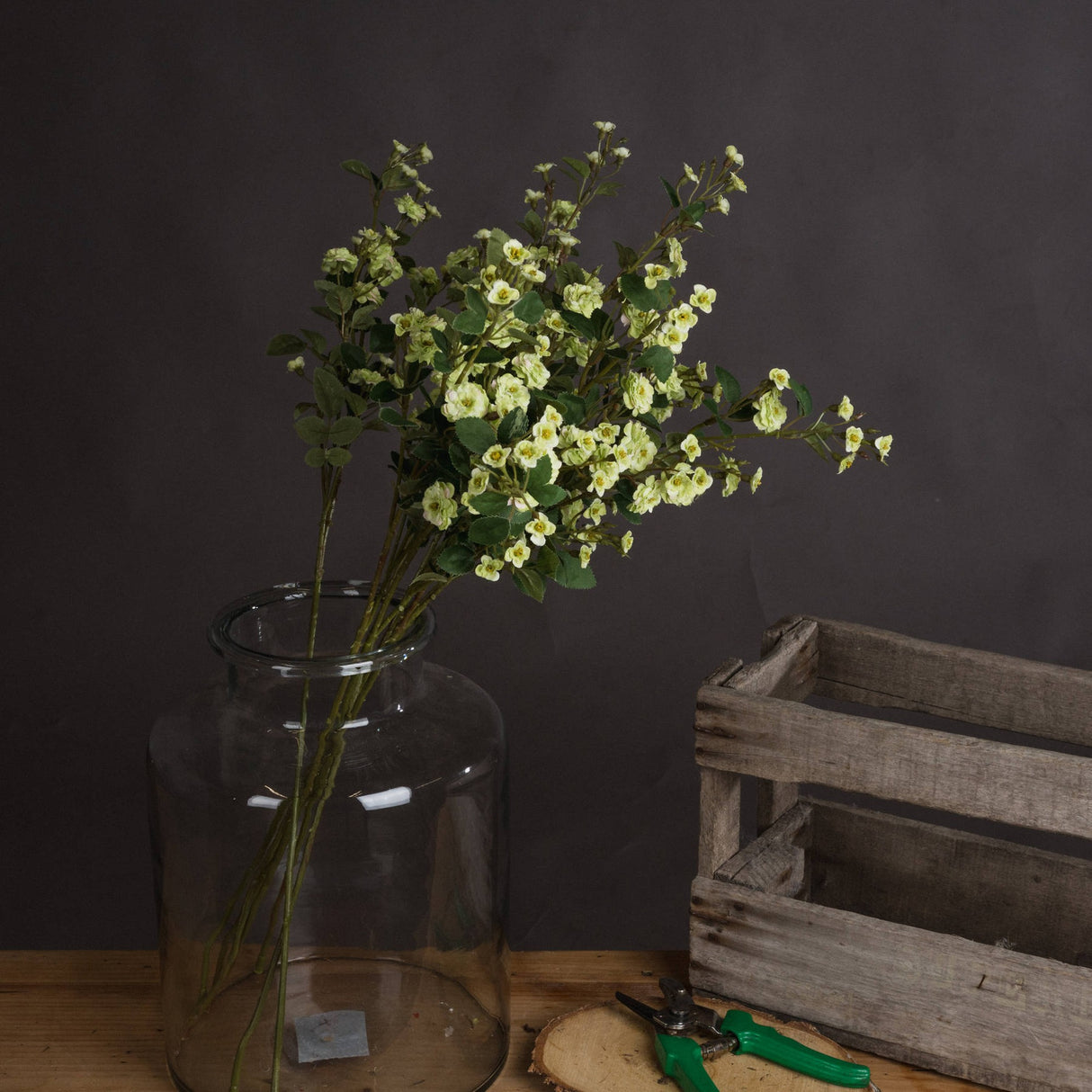 Hill Interiors Green Wild Meadow Rose faux stems with clustered pale green blooms and leaves in a tall glass jar vase beside a wooden crate.