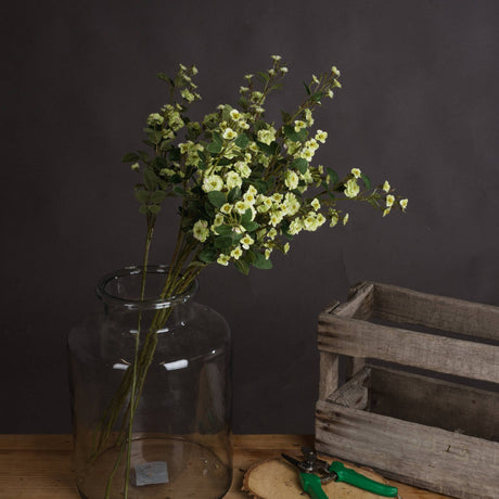 Hill Interiors Green Wild Meadow Rose faux stems with clustered pale green blooms and leaves in a tall glass jar vase beside a wooden crate.