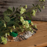 Hill Interiors Green Wild Meadow Rose faux stem with ruffled pale green blooms and veined leaves, resting on a birch wood slice against rustic timber.