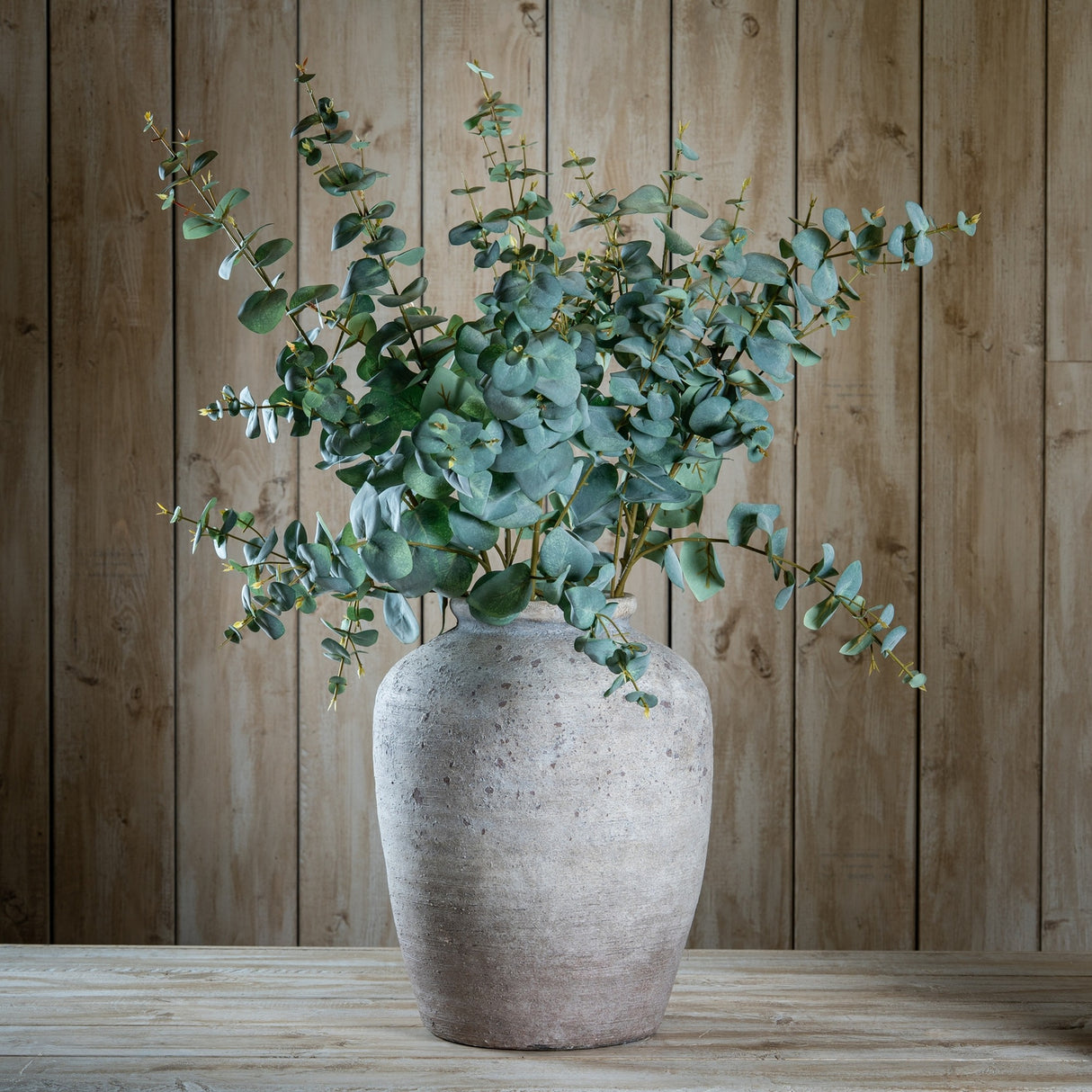 Hill Interiors Variegated Eucalyptus faux stems arranged in a rustic stone vase on a wooden table against a wooden plank wall.