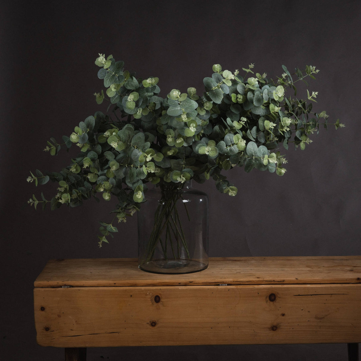Hill Interiors Eucalyptus Spray arrangement of faux sage-green foliage in a clear glass jar vase on a rustic wooden bench against a dark grey wall.
