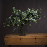 Hill Interiors Eucalyptus Spray arrangement of faux sage-green foliage in a clear glass jar vase on a rustic wooden bench against a dark grey wall.