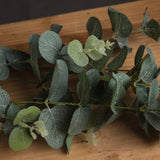 Hill Interiors Eucalyptus Spray with rounded sage green plastic leaves along slender stems, laid across a wooden surface.
