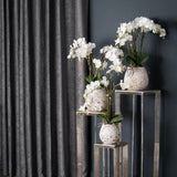 Hill Interiors White Orchid In Stone Pot display, three faux Phalaenopsis arrangements in chalky stone-effect pots on silver pedestals beside grey curtain.