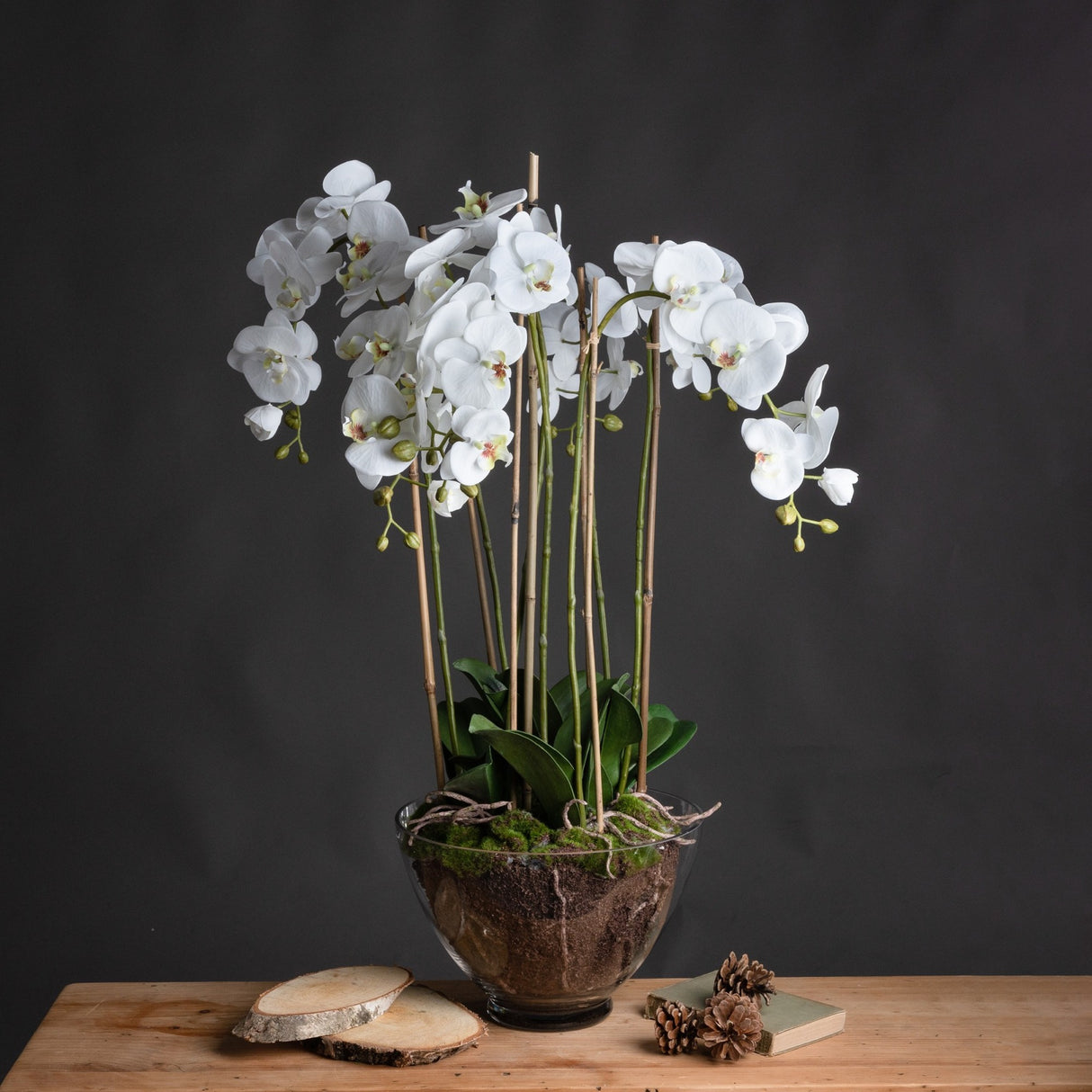 Large white faux Phalaenopsis orchid by Hill Interiors with arching stems and green leaves in a clear glass pot, styled on a wooden table.