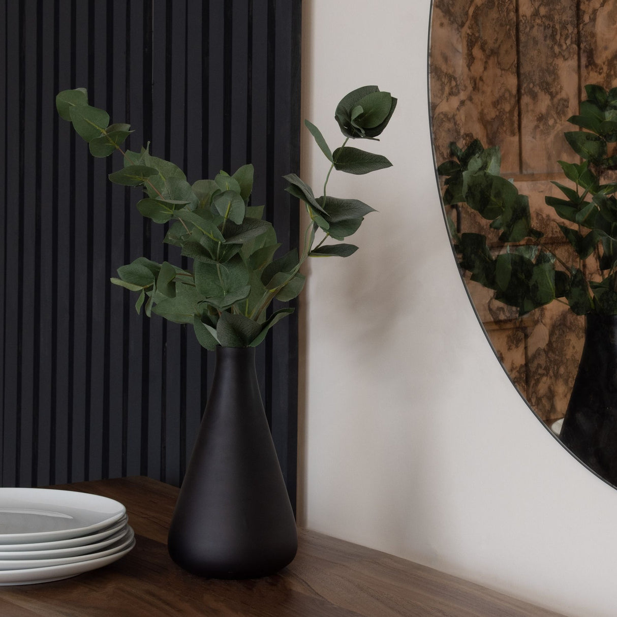 Hill Interiors Eucalyptus Filler stems arranged in a matte black tapered vase on a wooden sideboard beside white plates and a round mirror.
