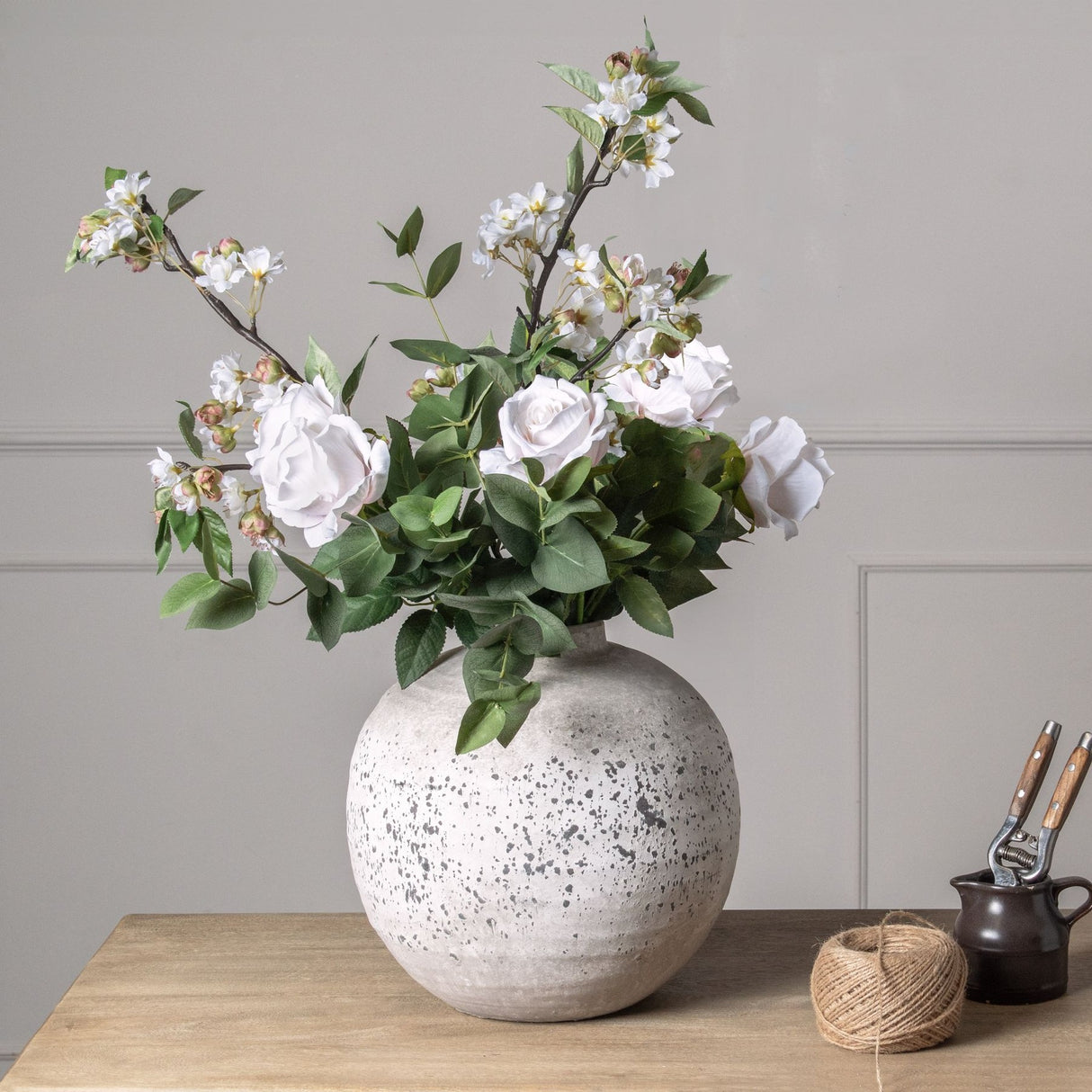 Hill Interiors Eucalyptus Filler arranged with white roses and blossom branches in a speckled stone ball vase on a wooden table.