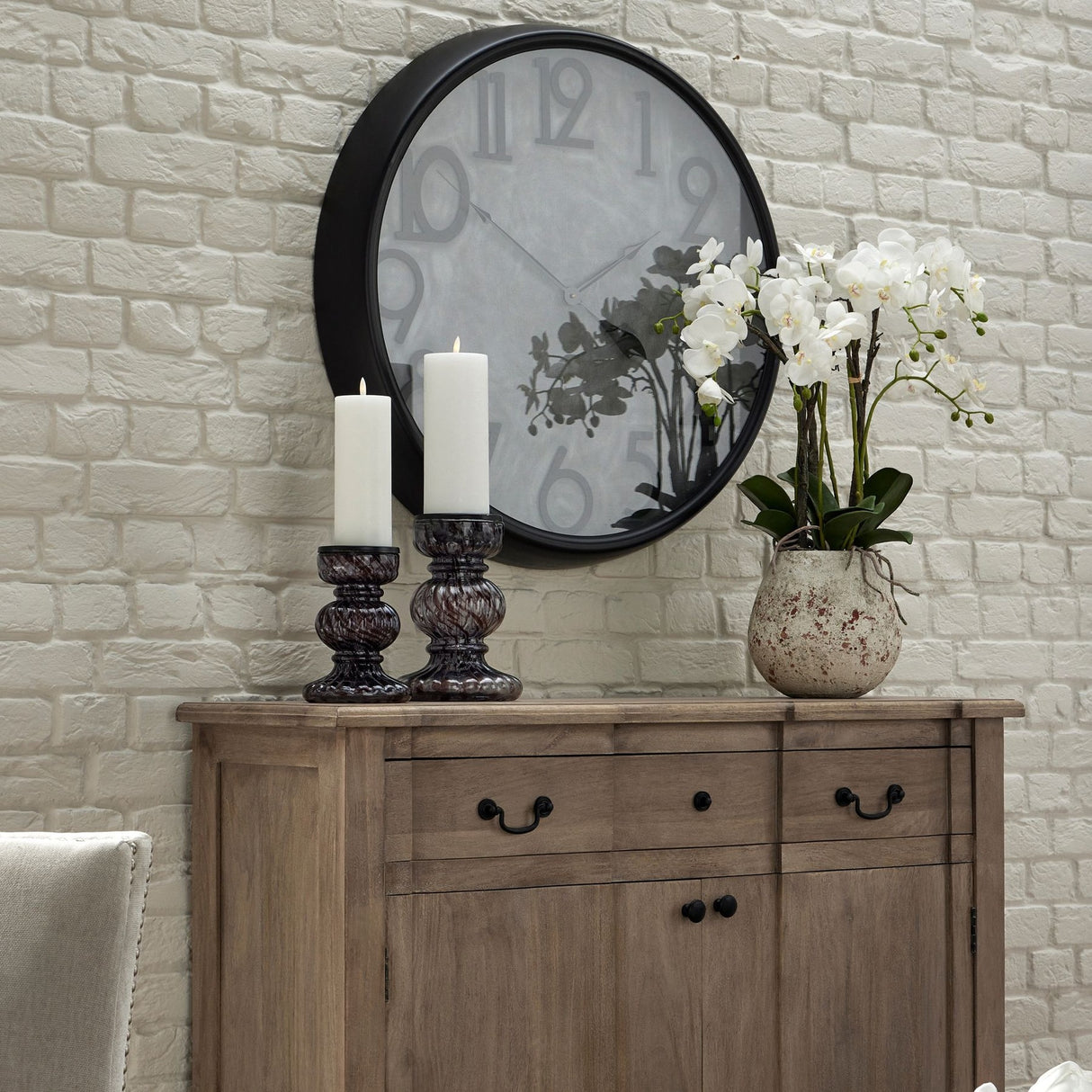 Hill Interiors Large Stone Potted Orchid With Roots displayed on a wooden sideboard beside candle holders, beneath a round black wall clock.