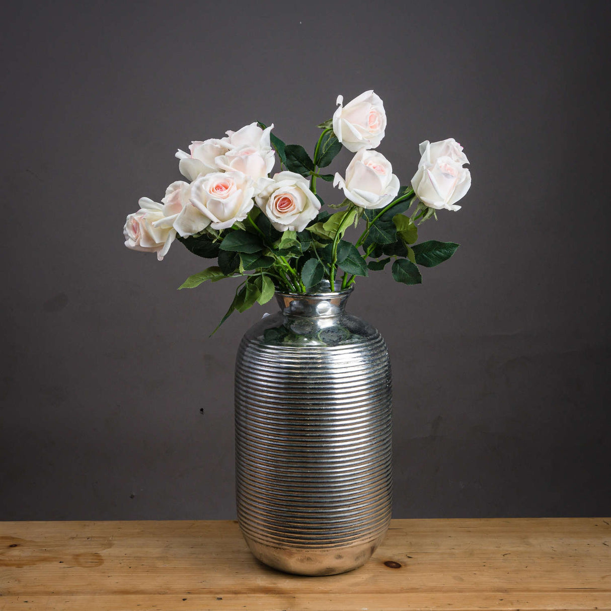 Hill Interiors Palest Pink Tea Rose faux blooms arranged in a ribbed silver vase on a wooden surface against a dark grey background.