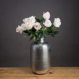Hill Interiors Palest Pink Tea Rose faux blooms arranged in a ribbed silver vase on a wooden surface against a dark grey background.
