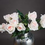 Hill Interiors Palest Pink Tea Rose faux stems arranged in a ribbed silver vase with green leaves, set against a dark grey background.