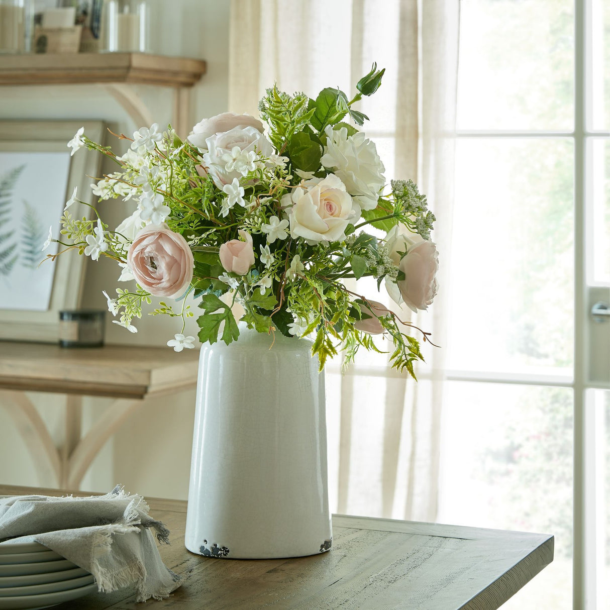 Hill Interiors Palest Pink Tea Rose arrangement with blush roses, ranunculus and white blooms in a crackle-glaze ceramic vase on a wooden table by a window.