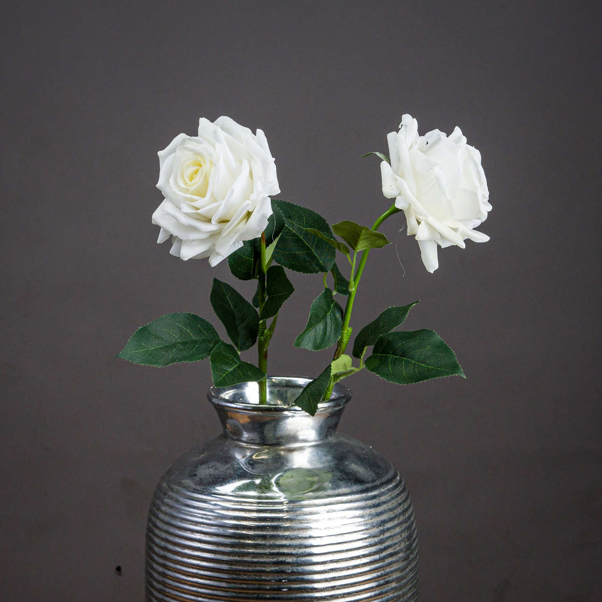 Large white garden rose faux stems by Hill Interiors in a ribbed silver vase, showing full blooms and green leaves against a grey backdrop.