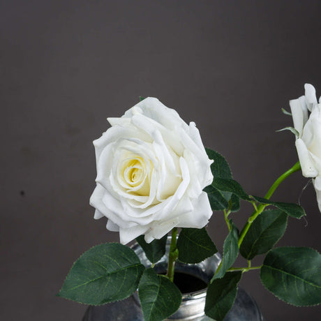 Hill Interiors Large White Garden Rose faux stem with layered petals and green leaves, displayed in a metal vessel against a grey background.